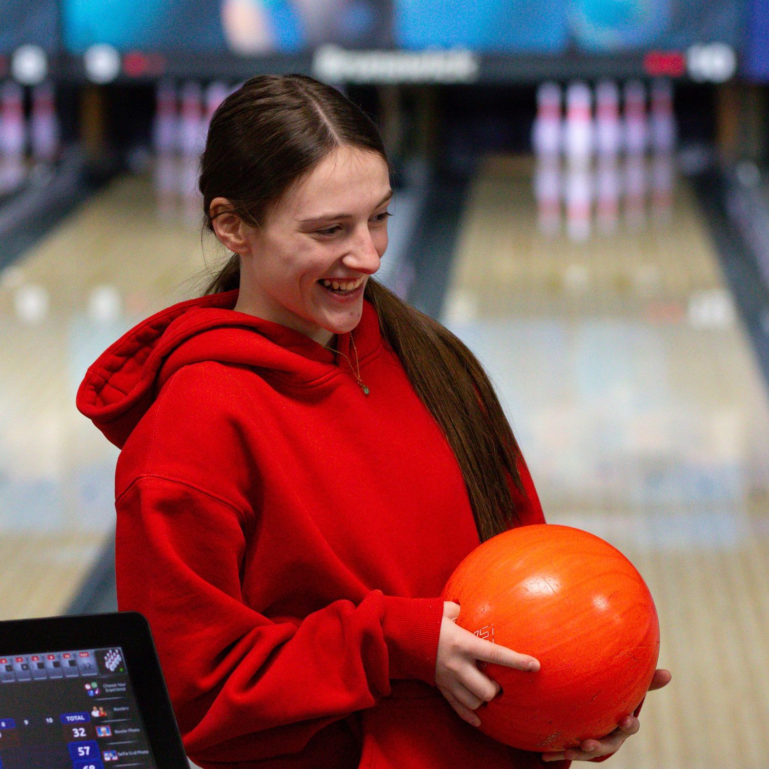 Smiling young woman in red hoodie holding an orange bowling ball at a bowling alley.