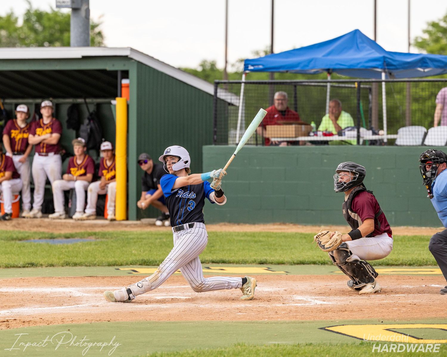 Youth baseball batter swings at pitch during game with catcher and dugout of players visible in background.