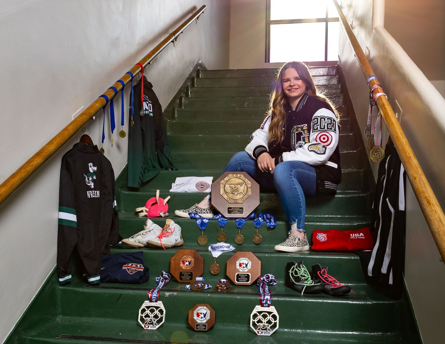 Young female athlete poses on green stairs surrounded by trophies, medals, and sports gear celebrating her achievements.