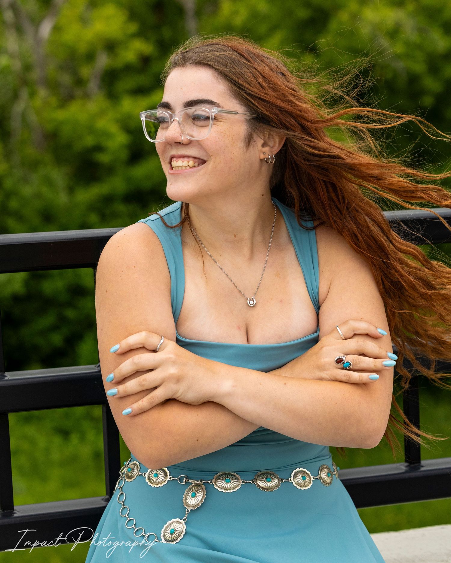 Young woman in teal dress with red hair blowing in wind, smiling outdoors on bridge, wearing glasses and jewelry.