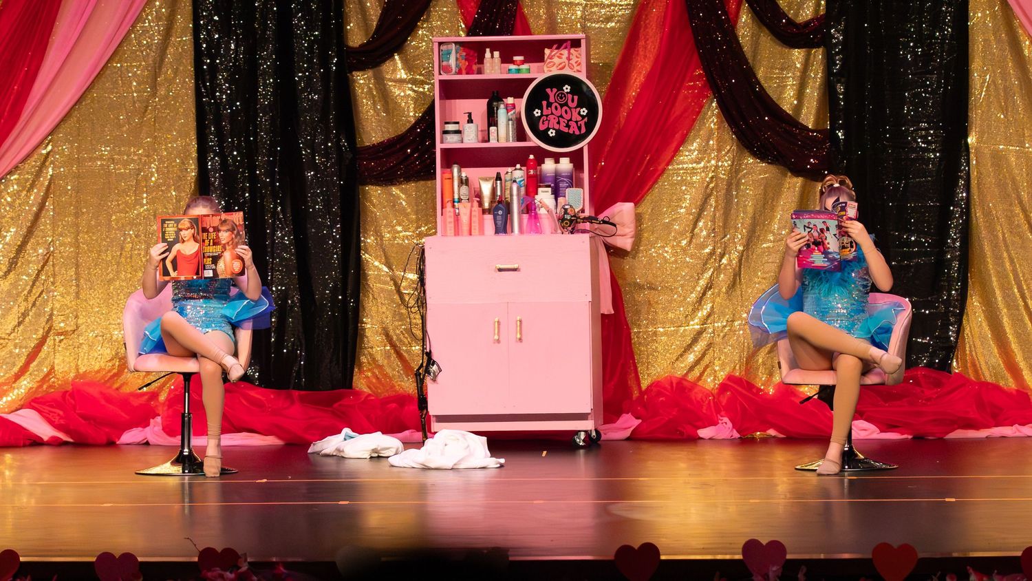 Two performers in colorful costumes pose on stage beside a pink cabinet during a vibrant dance recital performance.