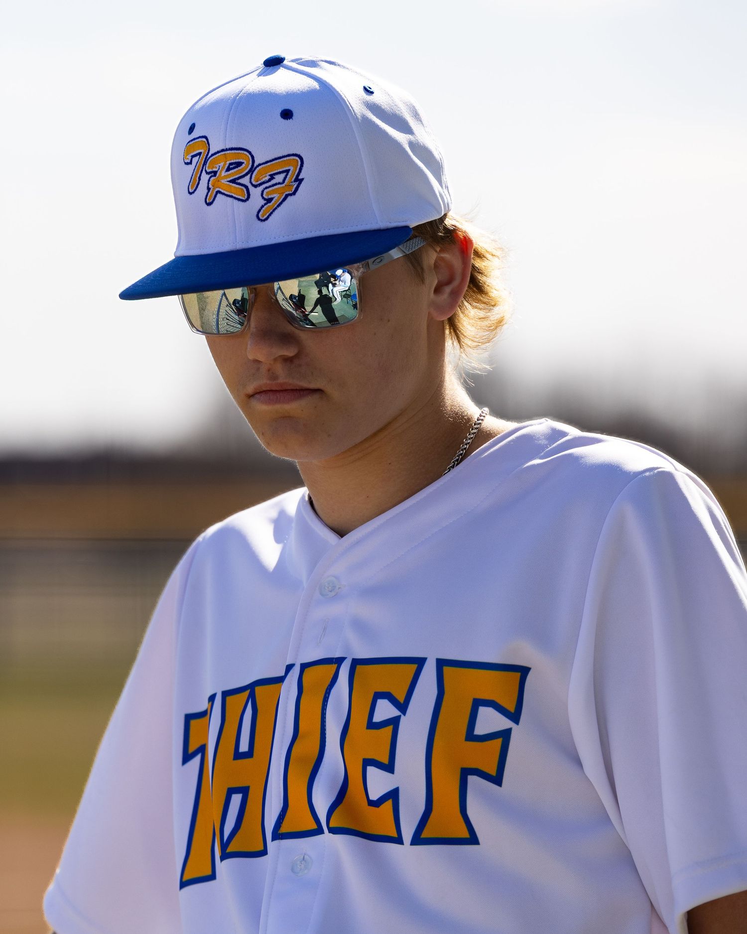 Young baseball player in white Thief jersey and blue TRF cap wearing mirrored sunglasses on field.