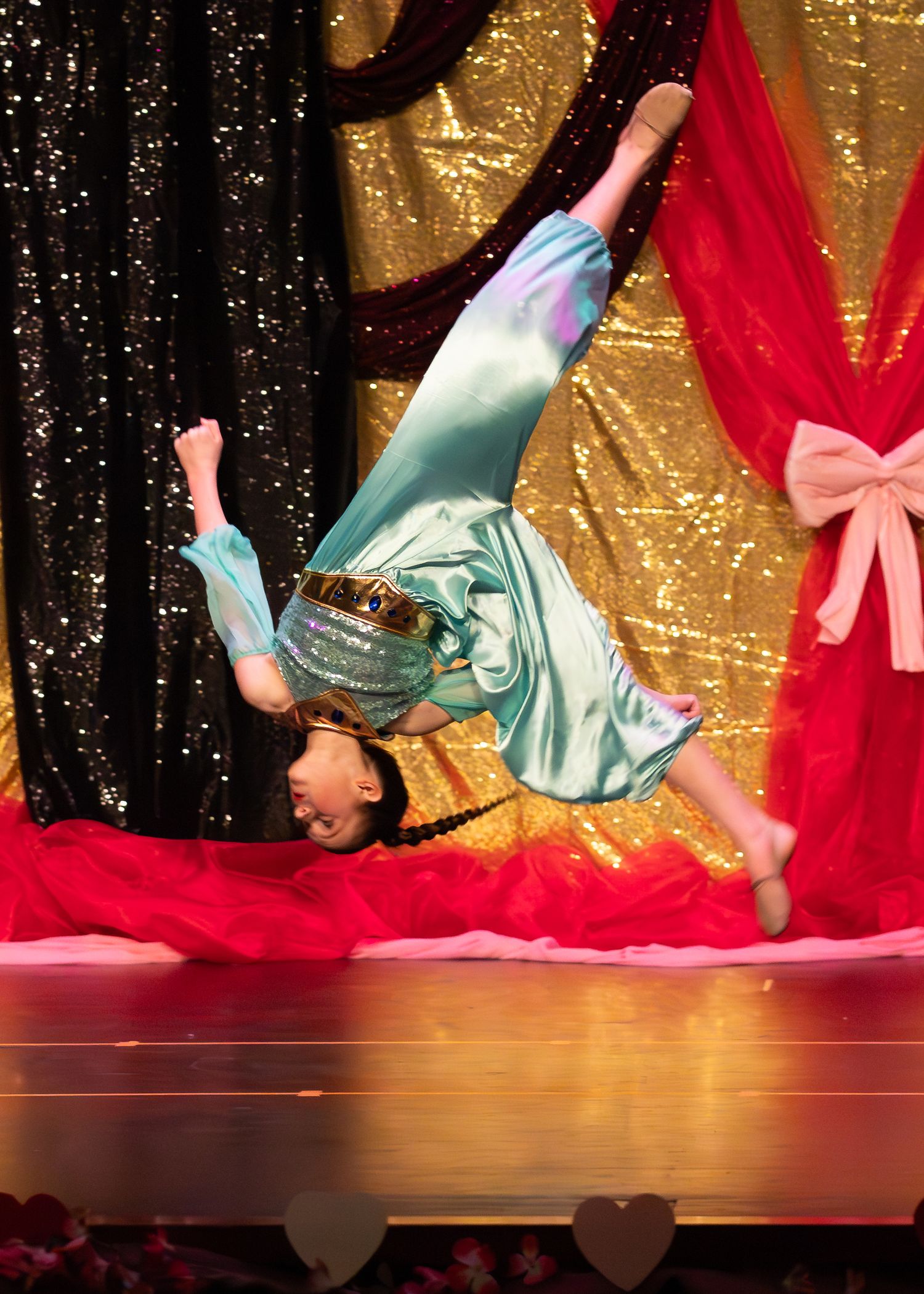 Dancer in teal satin costume executes an impressive backbend flip on a decorated performance stage.