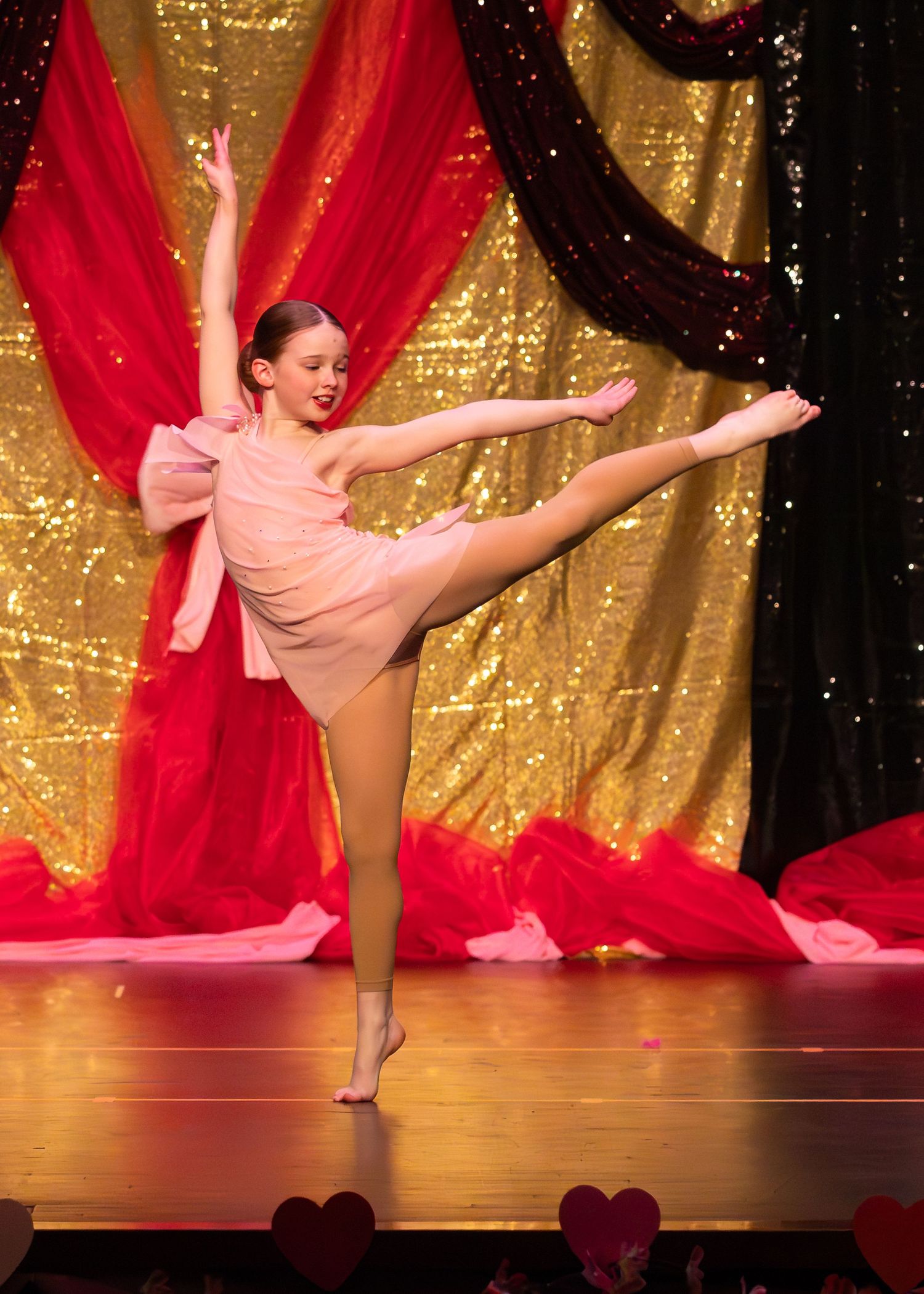 Young dancer performs arabesque on stage in pink costume with gold sequin backdrop and red draping.