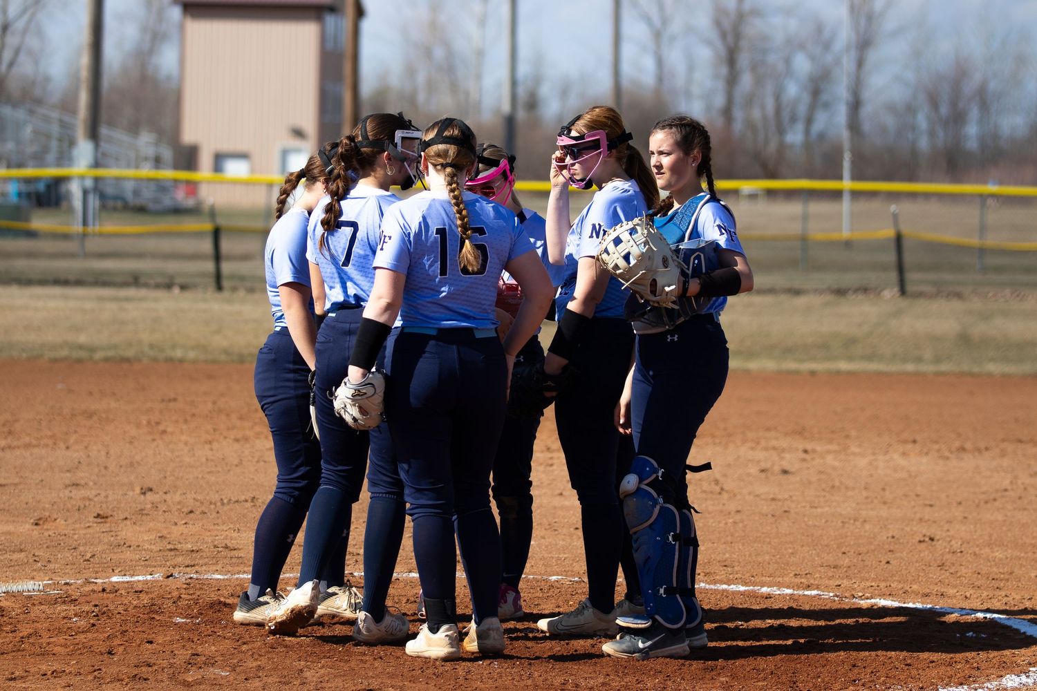 Girls softball team huddles on the pitcher's mound in blue uniforms during a spring game.