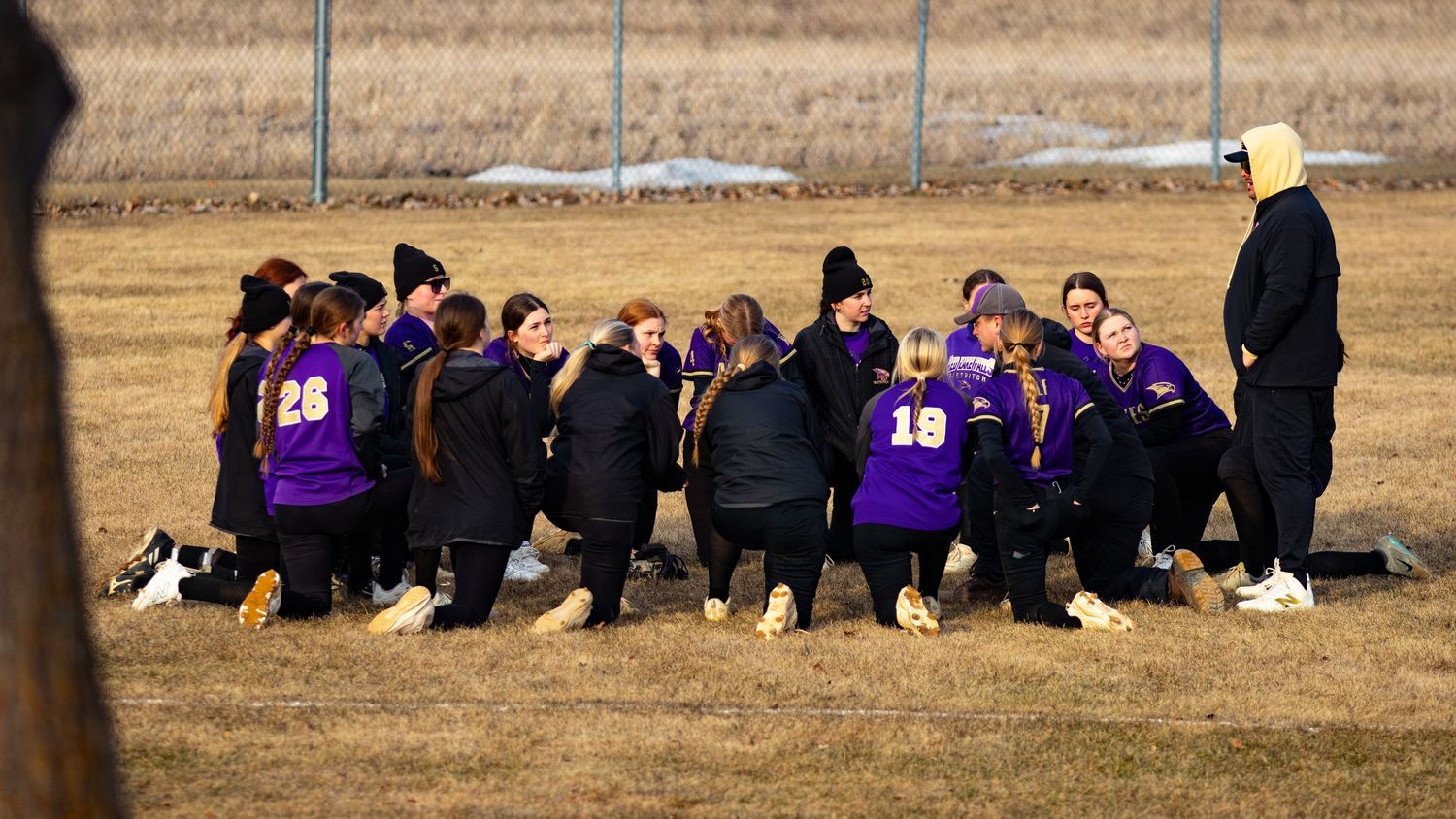 Youth softball team in purple jerseys huddles with coach on a field during cold weather practice.