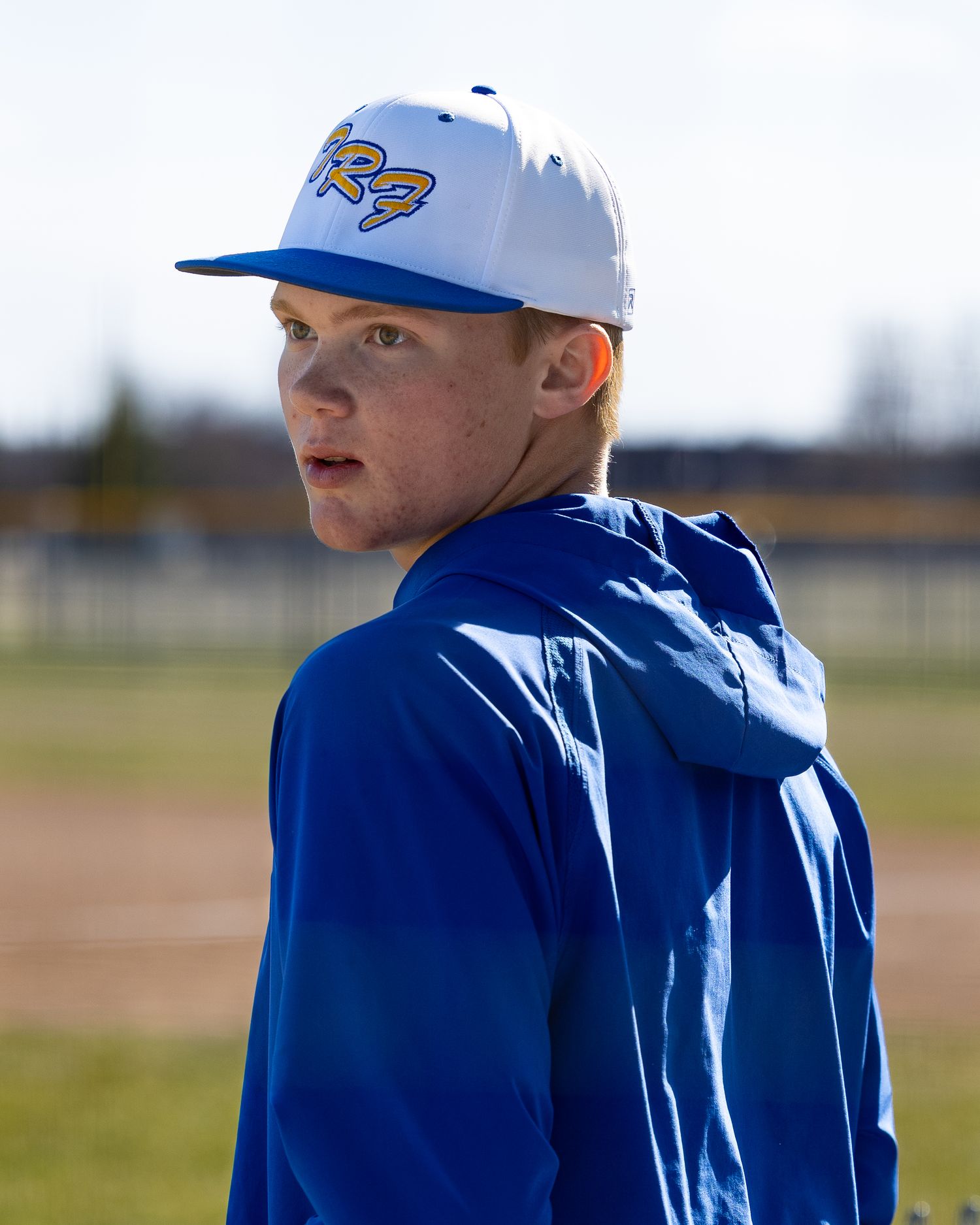 Young male baseball player in blue jacket and white cap with logo looks over shoulder on field.