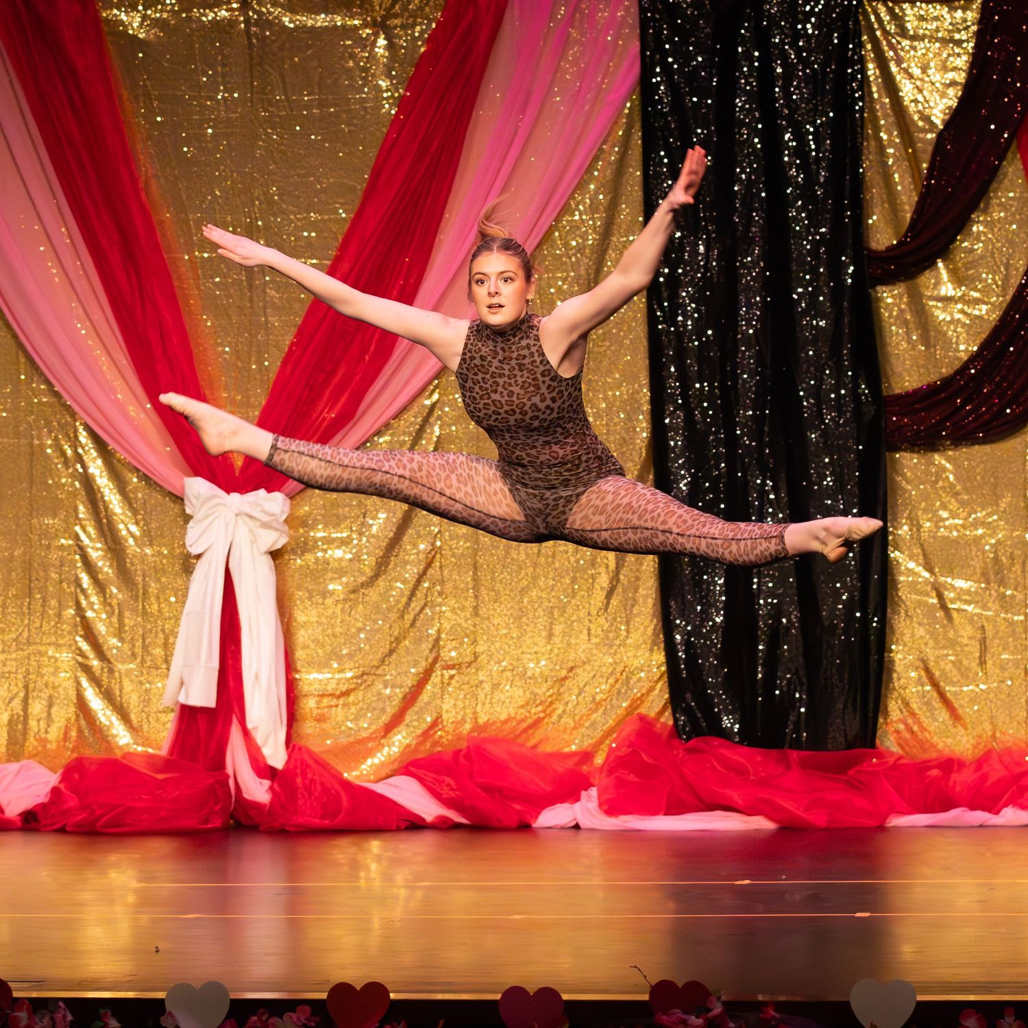Dancer in sparkly bodysuit performs a perfect split leap on stage with red and gold sequin backdrop.