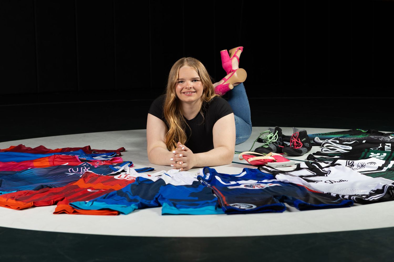 Smiling woman lying on floor surrounded by colorful sports jerseys in red, blue, and black on white circle.