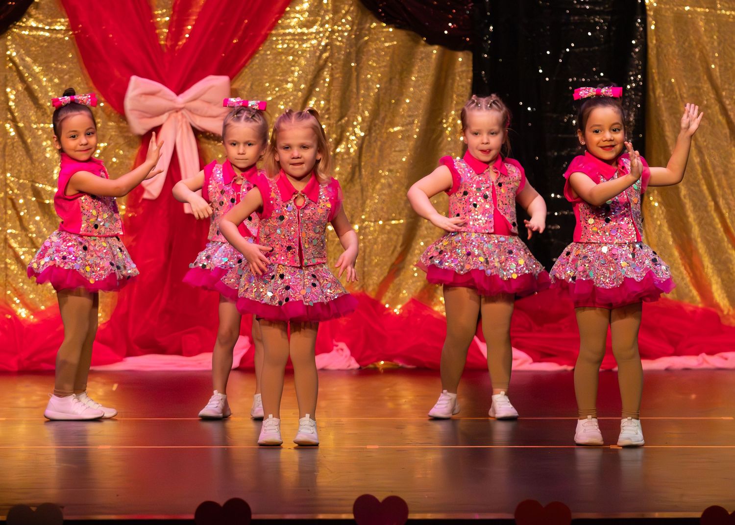 Five young girls in matching pink costumes perform a dance recital on a glittery stage.