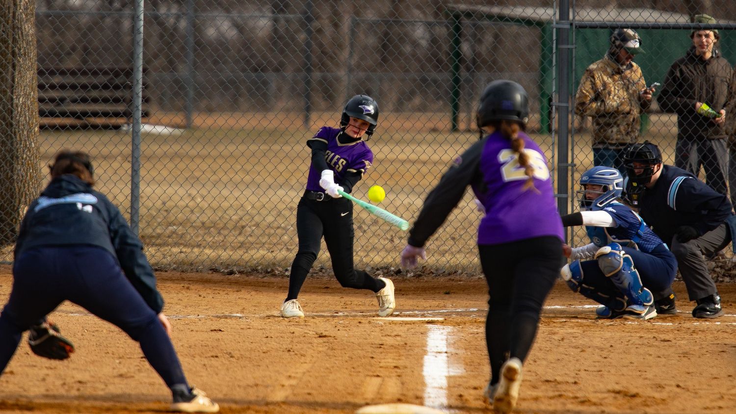 Girls softball player in purple uniform swings at a pitch during a youth league game on an outdoor diamond.