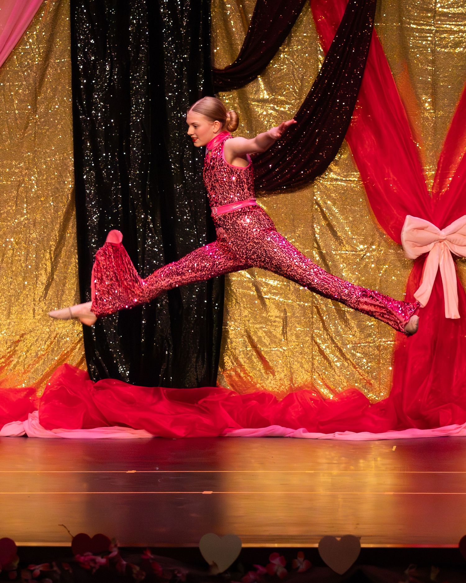 Young dancer in pink sequin jumpsuit performs a stunning leap on stage with gold and black sequin backdrop.