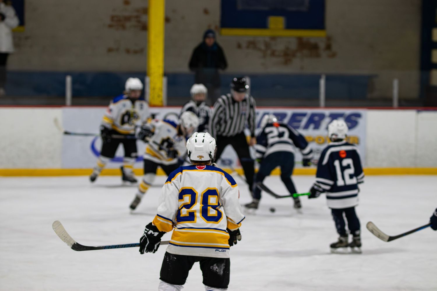 Youth hockey player wearing number 26 watches teammates compete during indoor ice hockey game.