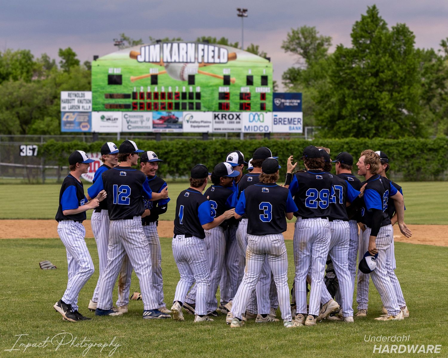 Baseball team celebrating on Jim Karn Field, players in blue and white uniforms huddle together after a victory.