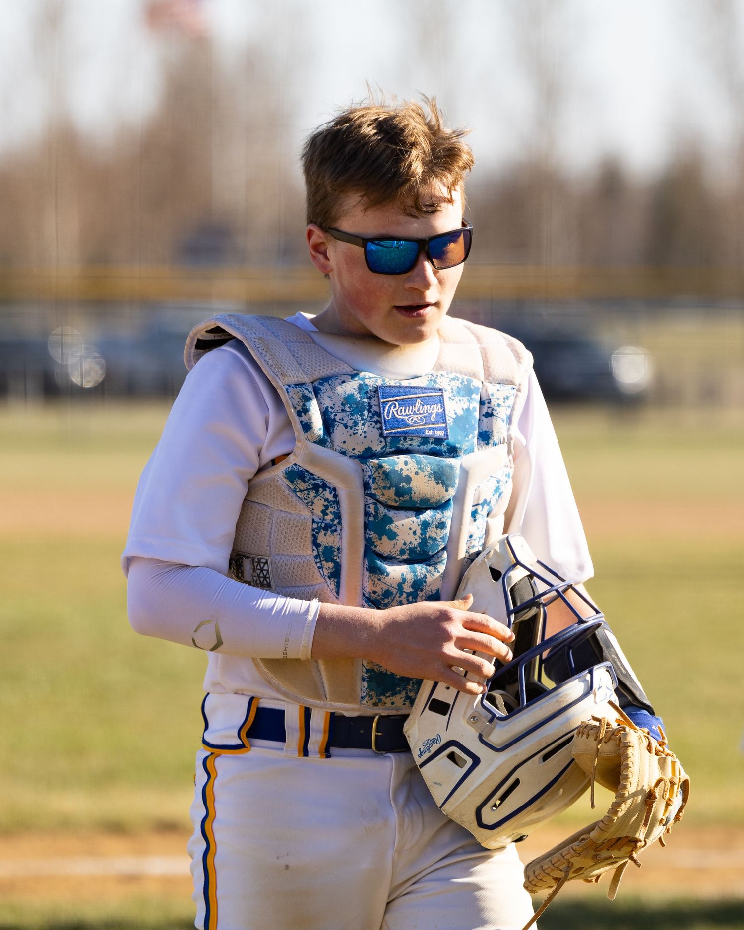 Young baseball catcher in Rawlings chest protector and sunglasses holding mitt on sunny field.
