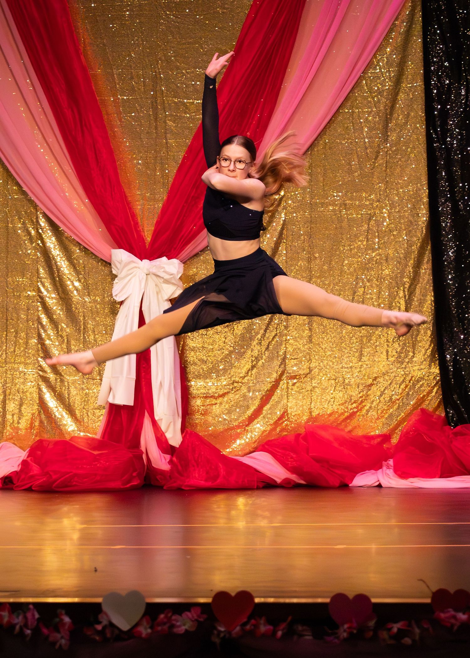 Dancer in black outfit performs dramatic leap split on stage with gold sequin and red fabric backdrop.