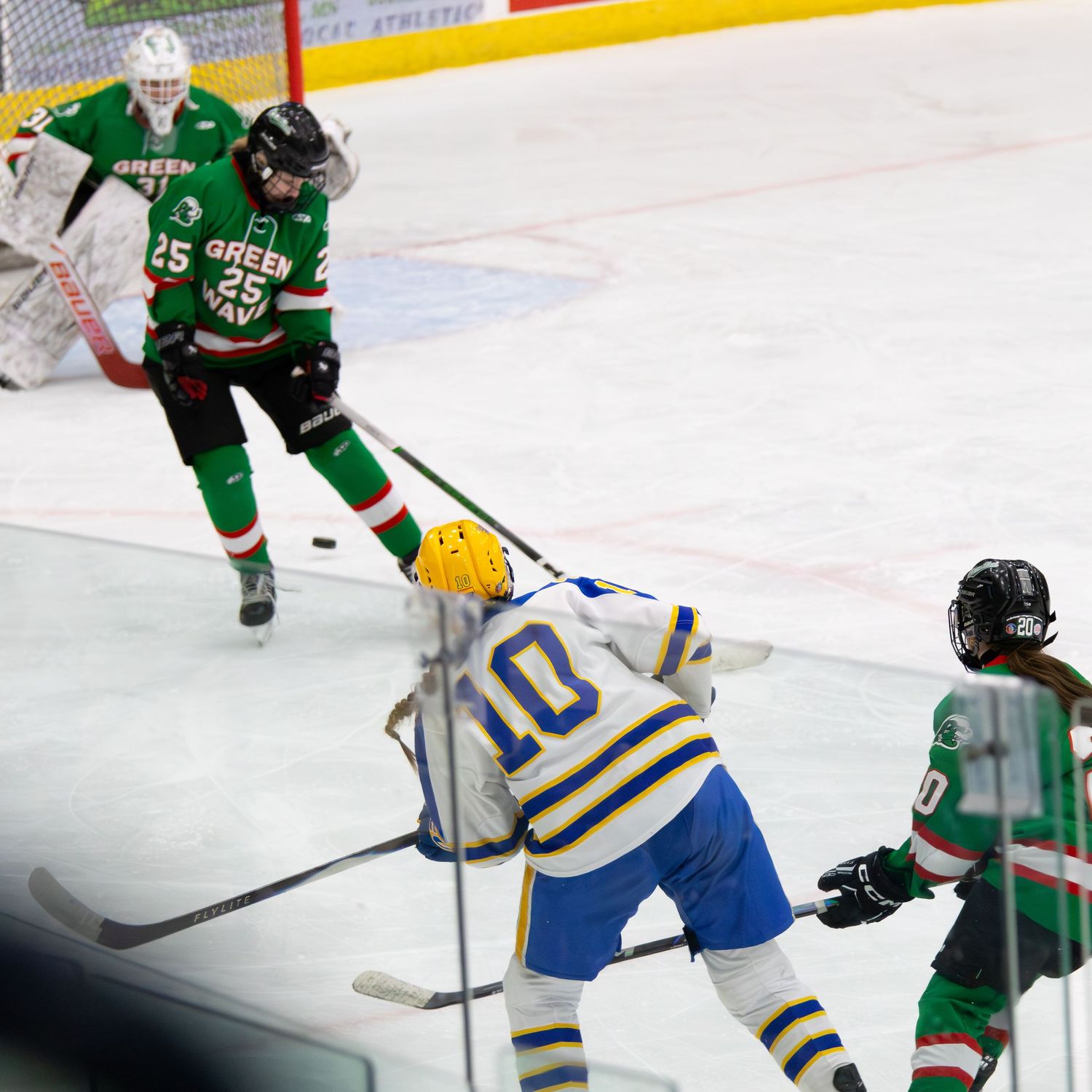Girls hockey players in blue and green jerseys compete near the goal.