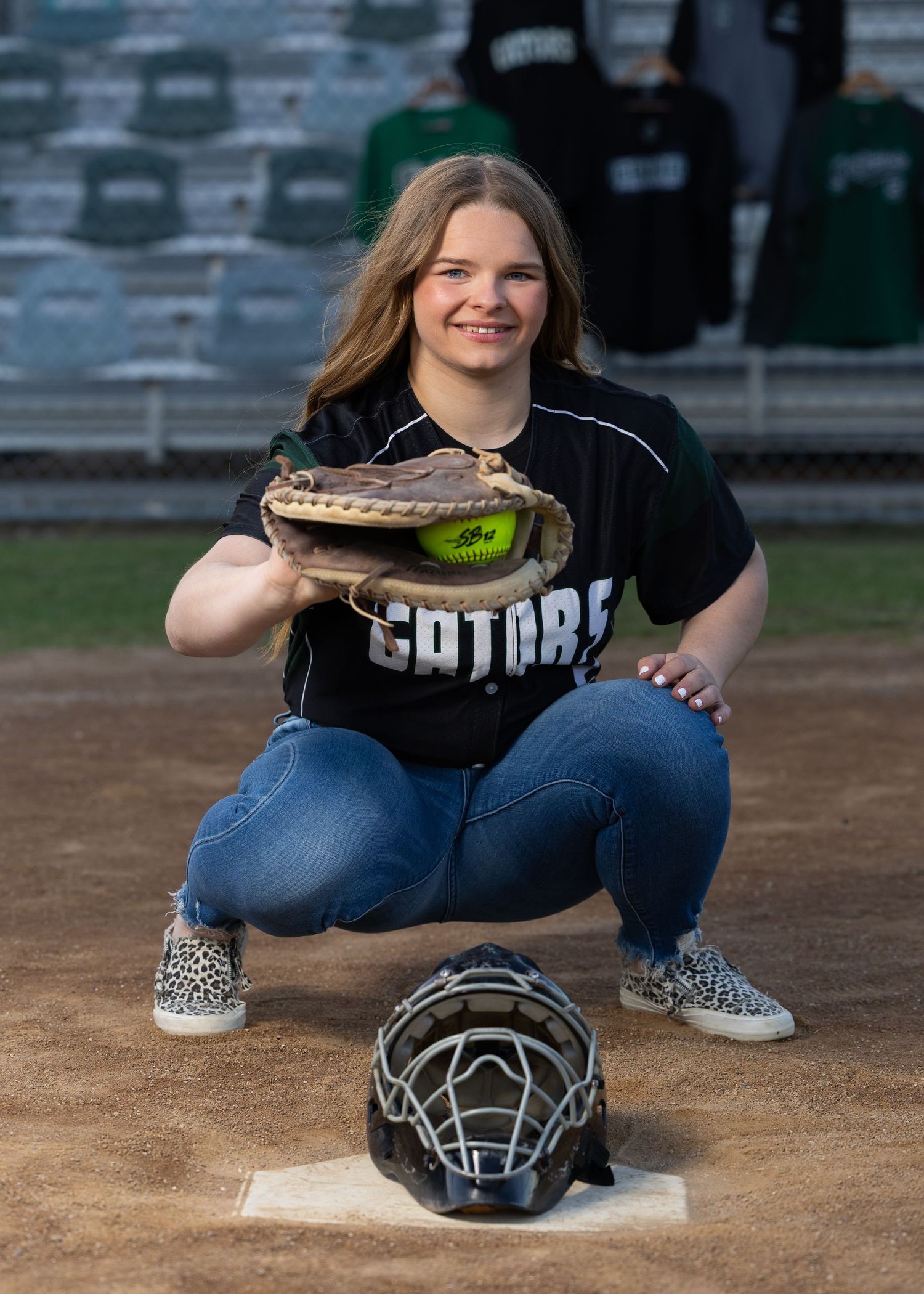 Young softball catcher in Gators jersey kneeling at home plate, holding glove with ball, helmet on ground.