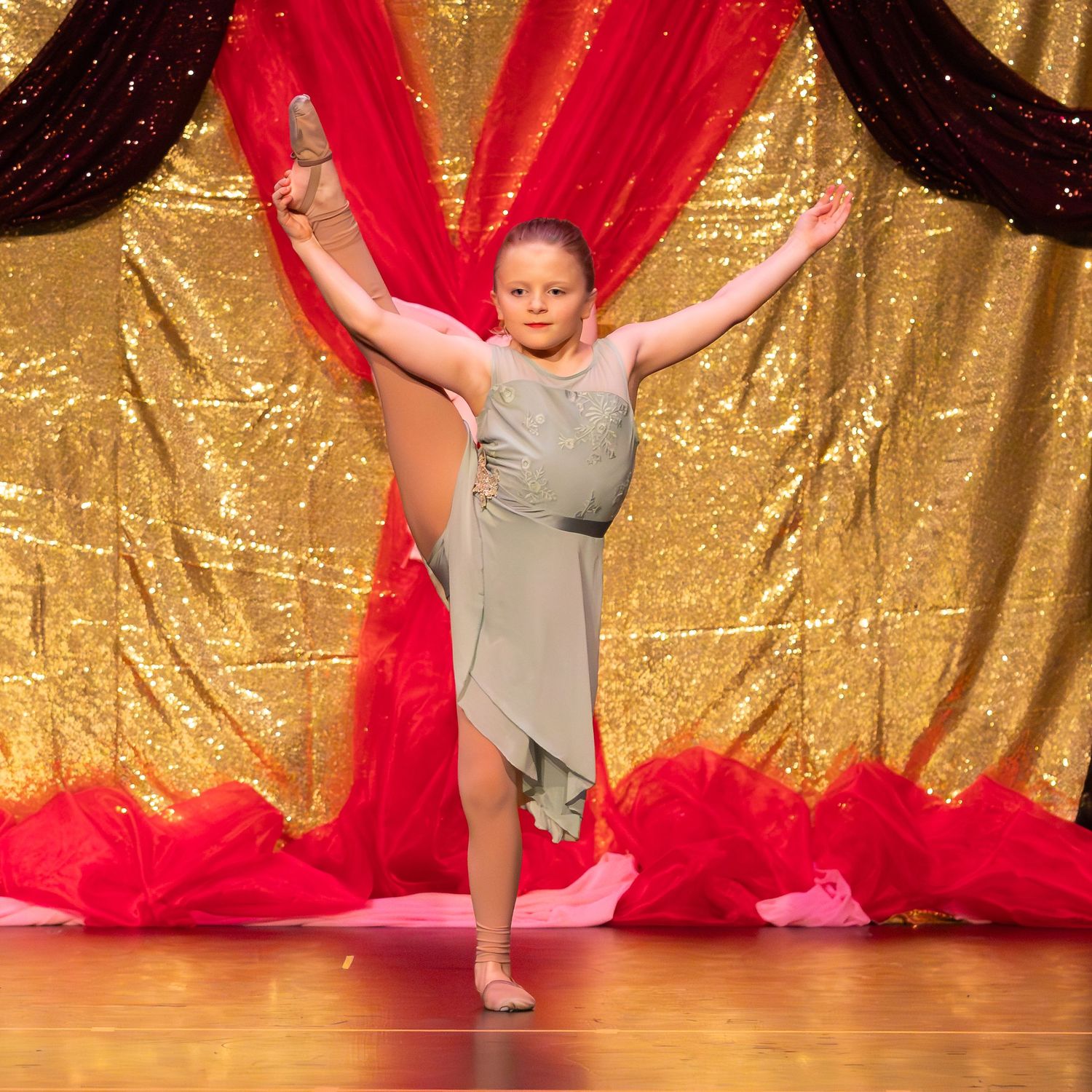 Young girl in gray dress performing a high kick on a glittery gold stage backdrop.