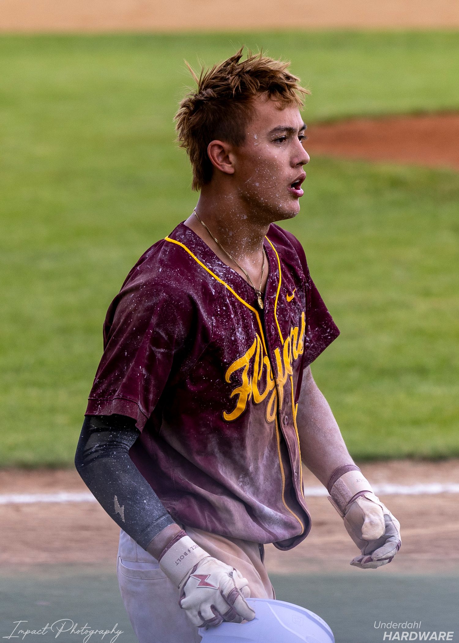 Dirt-covered baseball player in maroon Flynn jersey reacts after sliding, wearing batting gloves on the field.