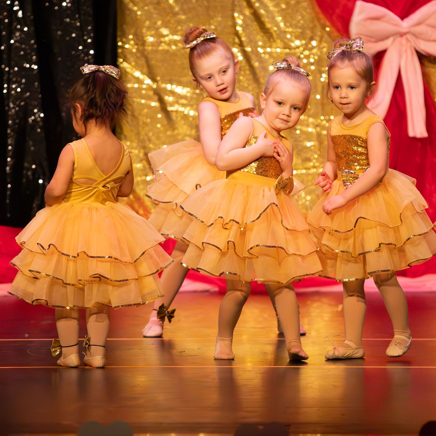 Four little girls in gold tutu dresses performing on stage at a children's dance recital.