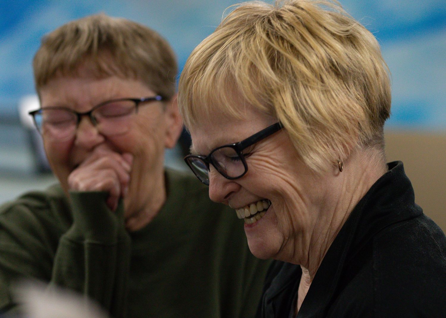 Two older women laughing together, one wearing glasses, in a bright indoor setting.