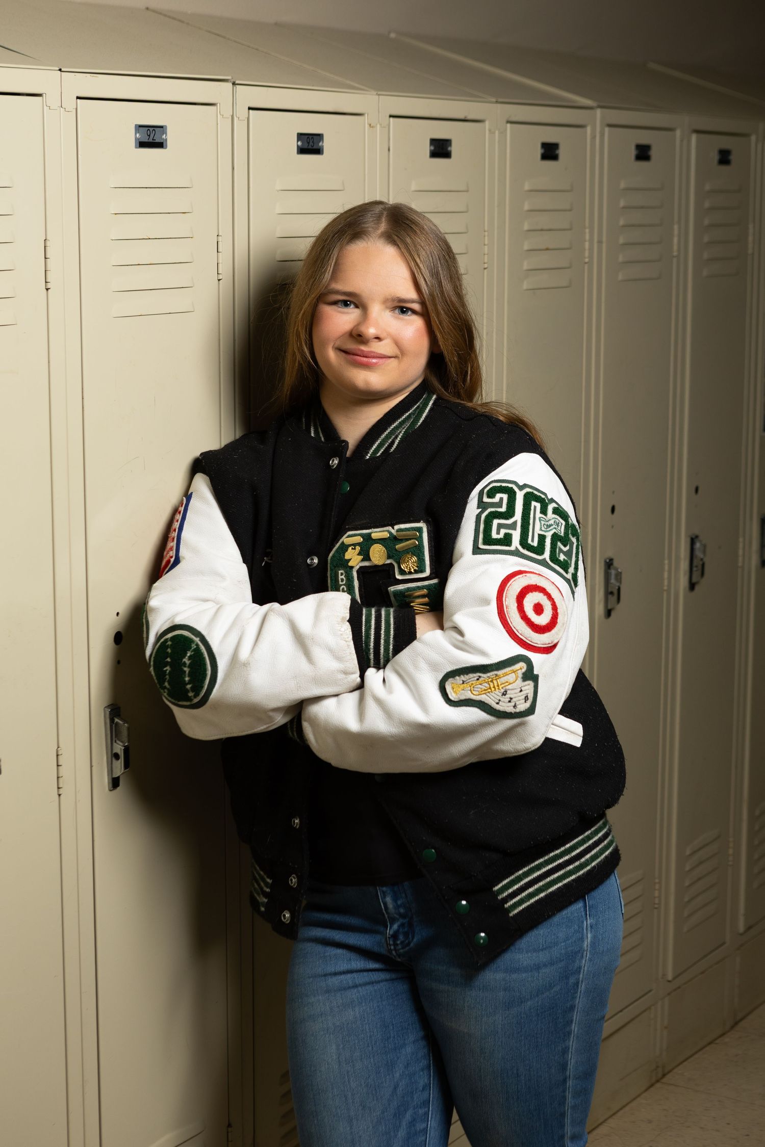 Teen girl in varsity letterman jacket leaning against school lockers with arms crossed, smiling confidently.
