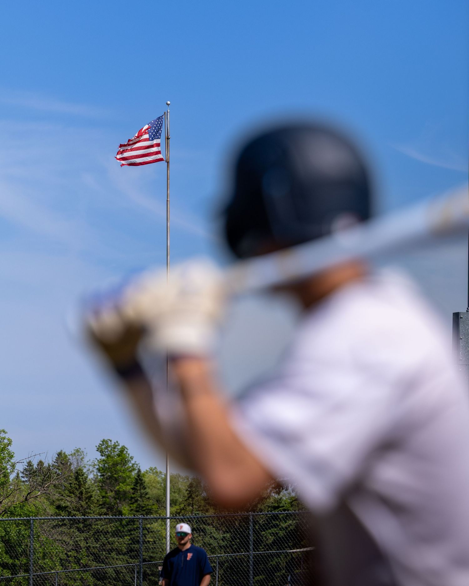 Baseball batter in helmet readies at plate with American flag flying on pole under clear blue sky.
