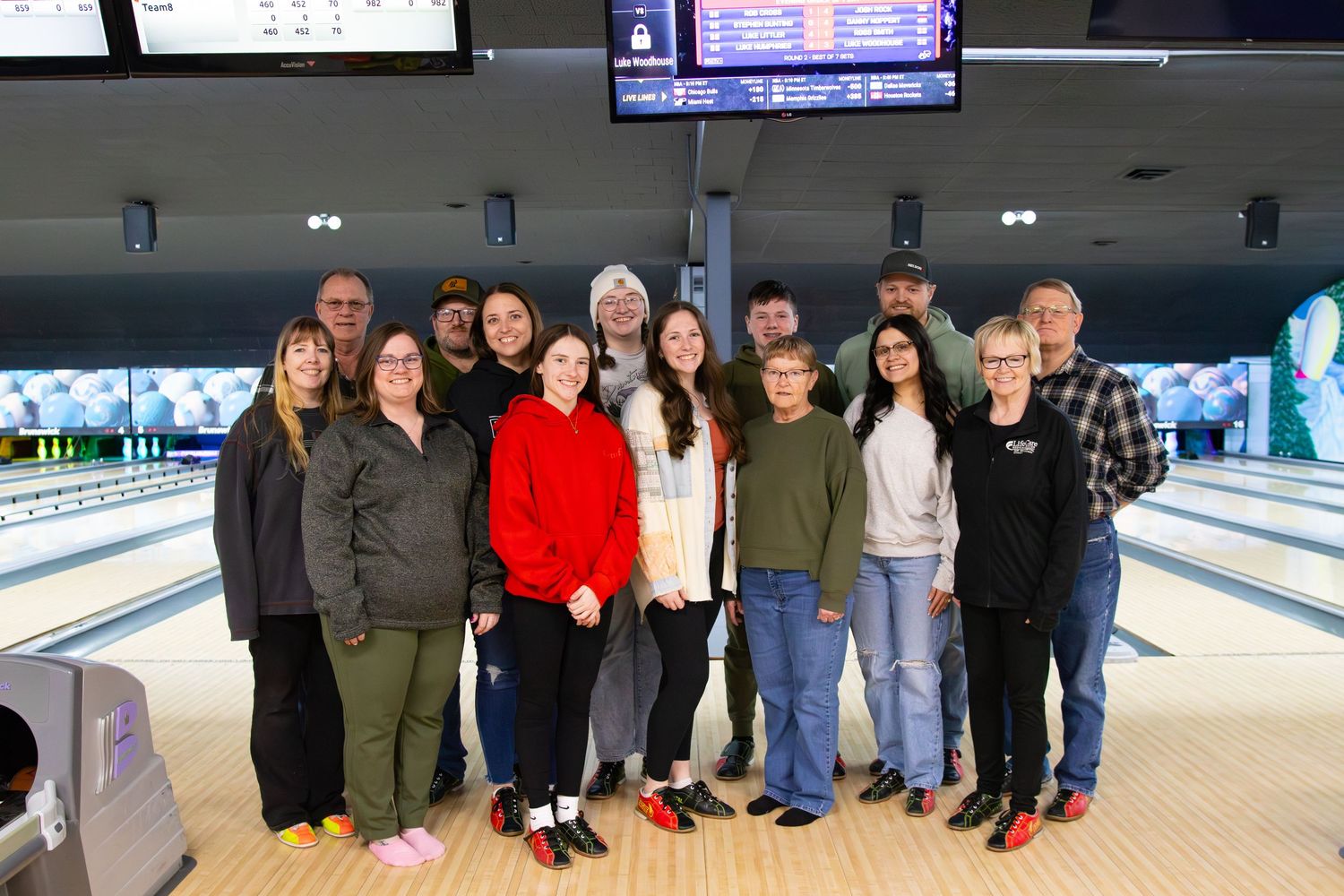 Group of people posing together in a bowling alley under a scoring screen.
