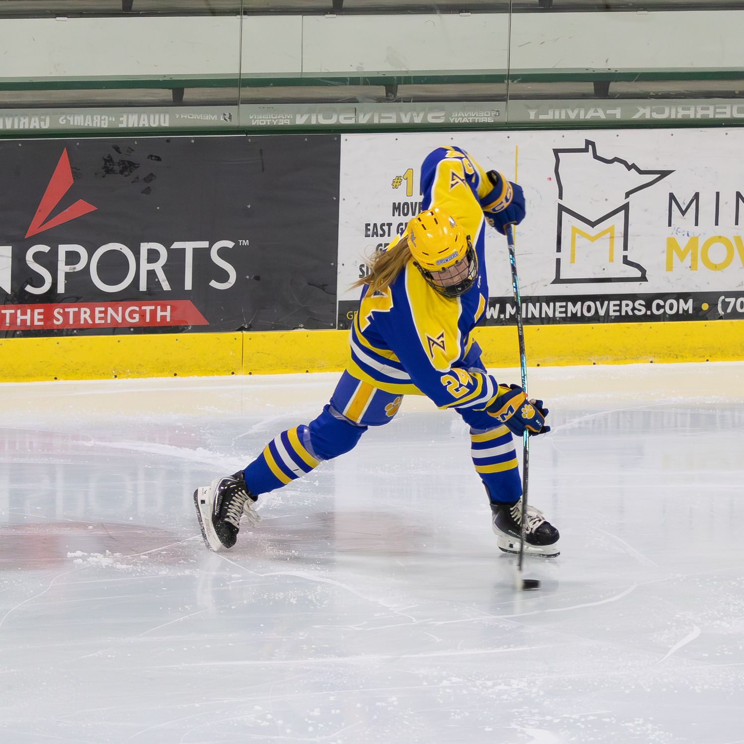 Youth hockey player in blue and yellow uniform takes a slap shot on an indoor ice rink near the boards. Stick bend.