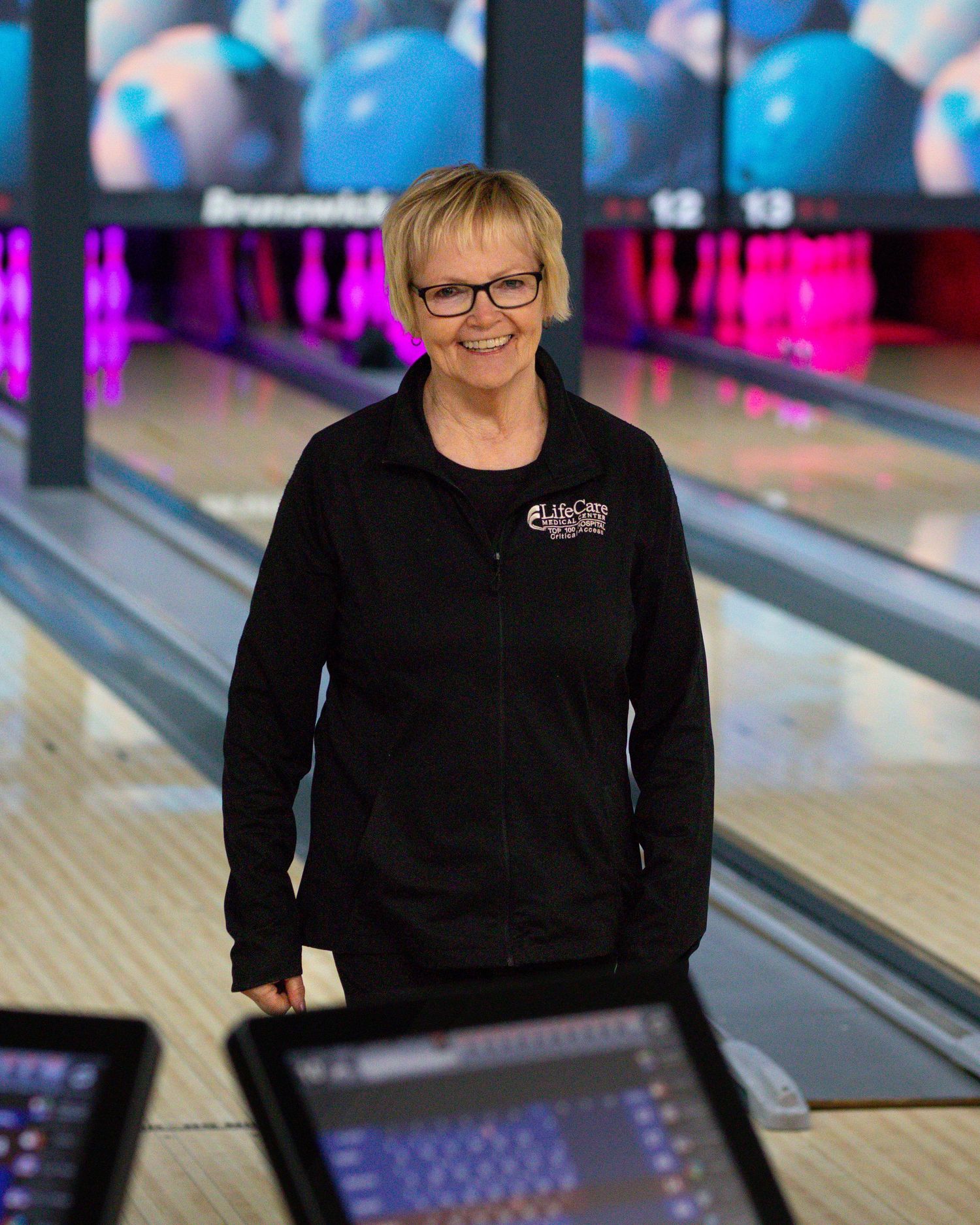 A smiling woman in a black jacket stands in a bowling alley with lanes in the background.