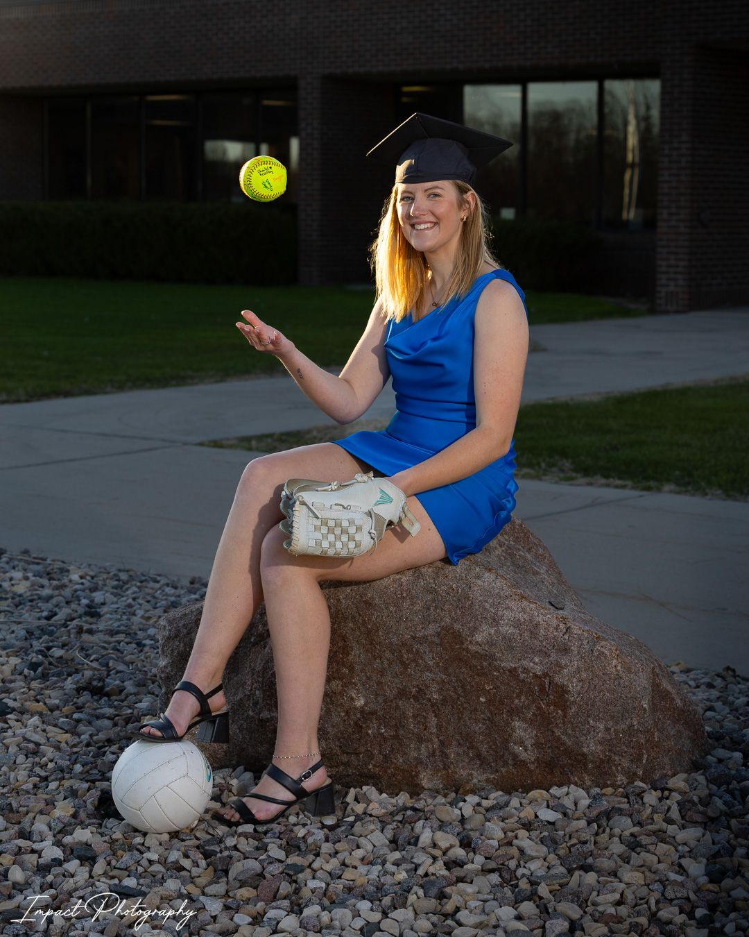 Young woman in blue dress and graduation cap sits on rock, tossing softball, with volleyball and glove nearby.