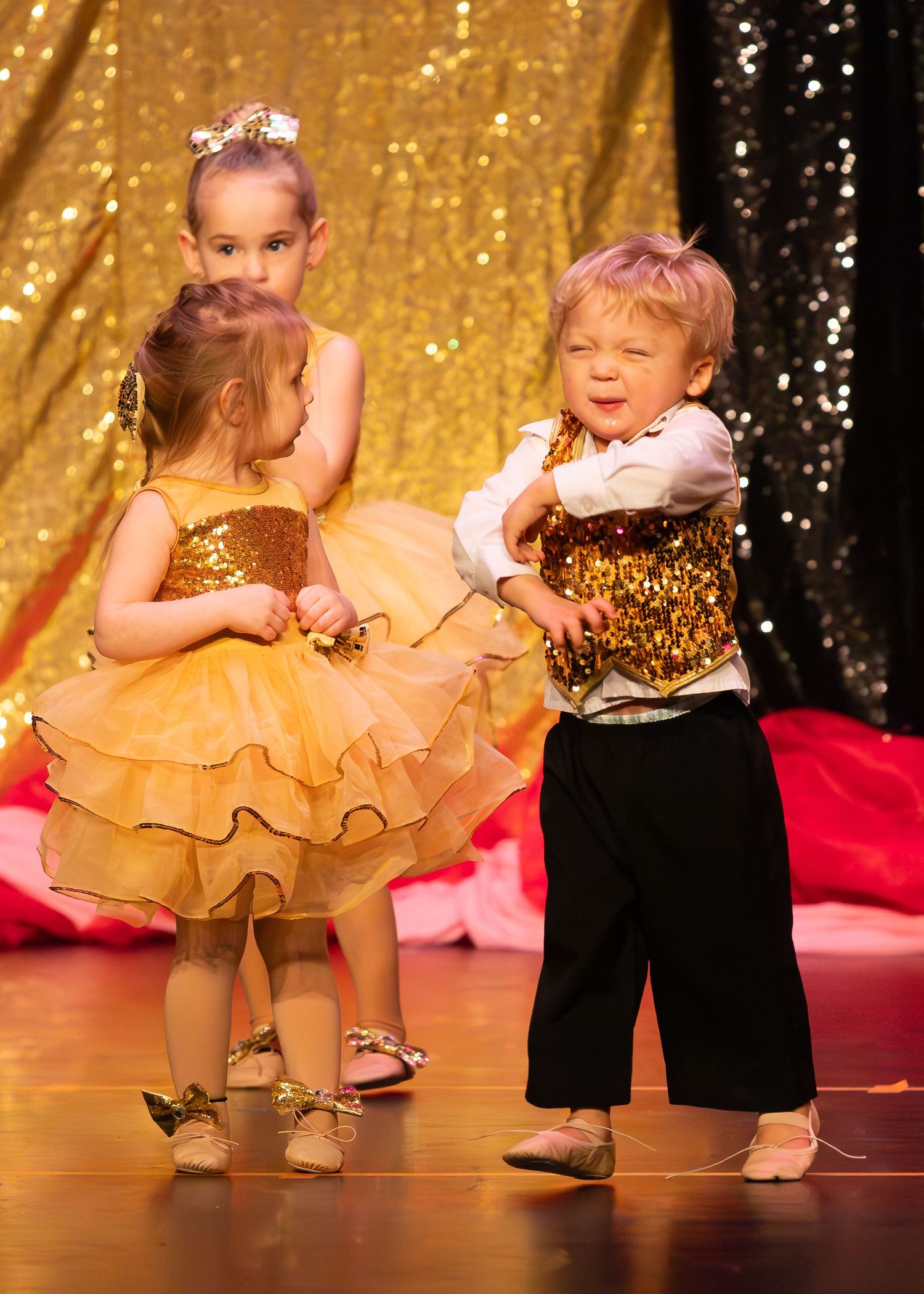 Three toddlers in gold sequin costumes perform on stage during a dance recital.