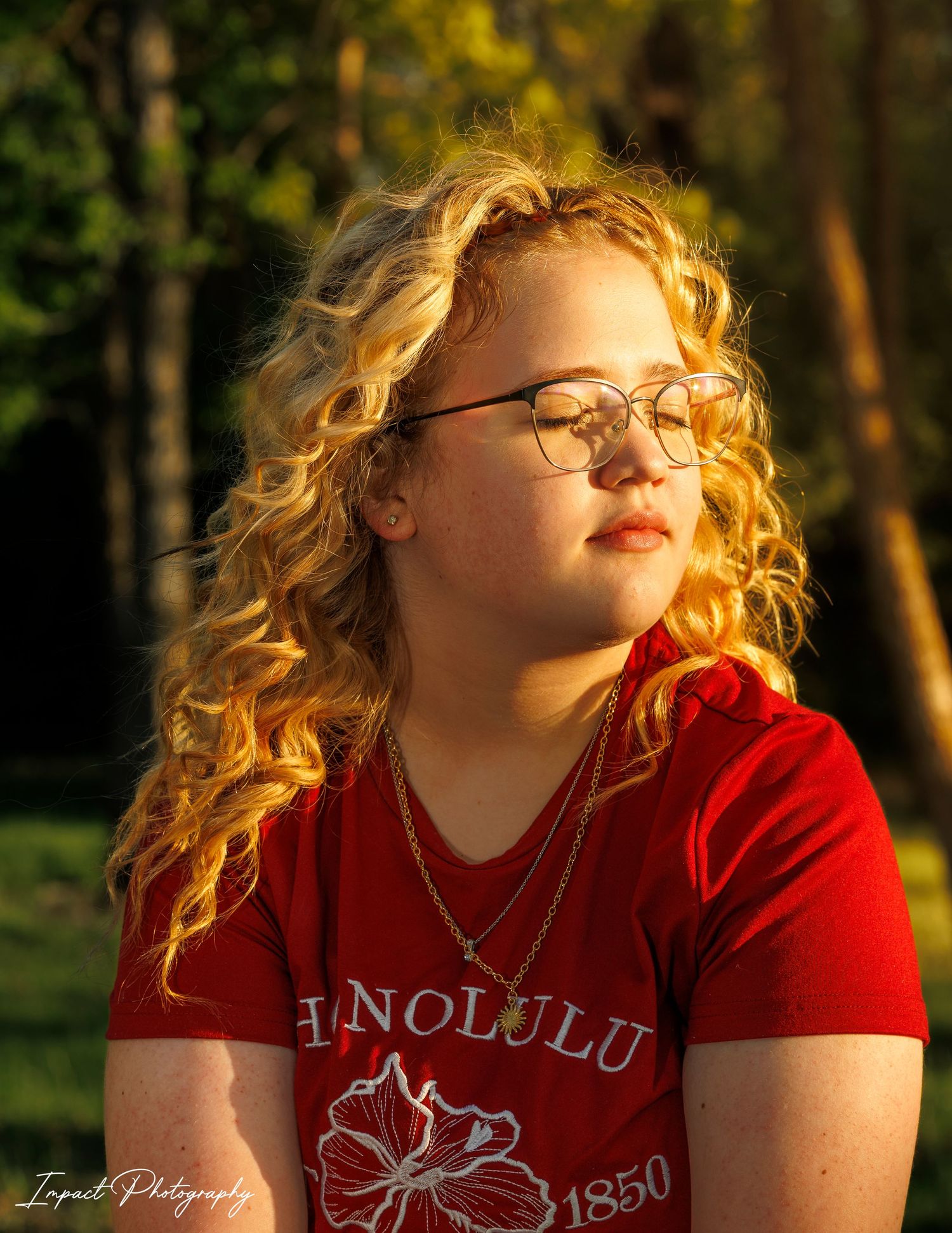 Young girl with curly blonde hair and glasses wearing a red Honolulu shirt, bathed in warm golden sunlight outdoors.