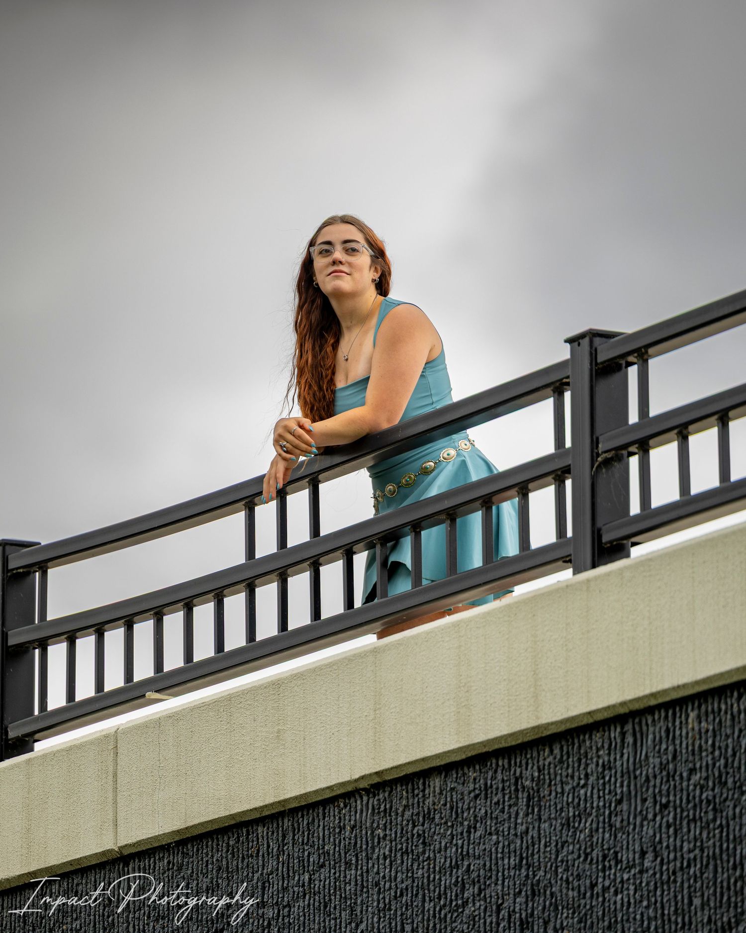 Young woman in teal dress leaning on dark railing of elevated balcony against cloudy sky, Impact Photography.