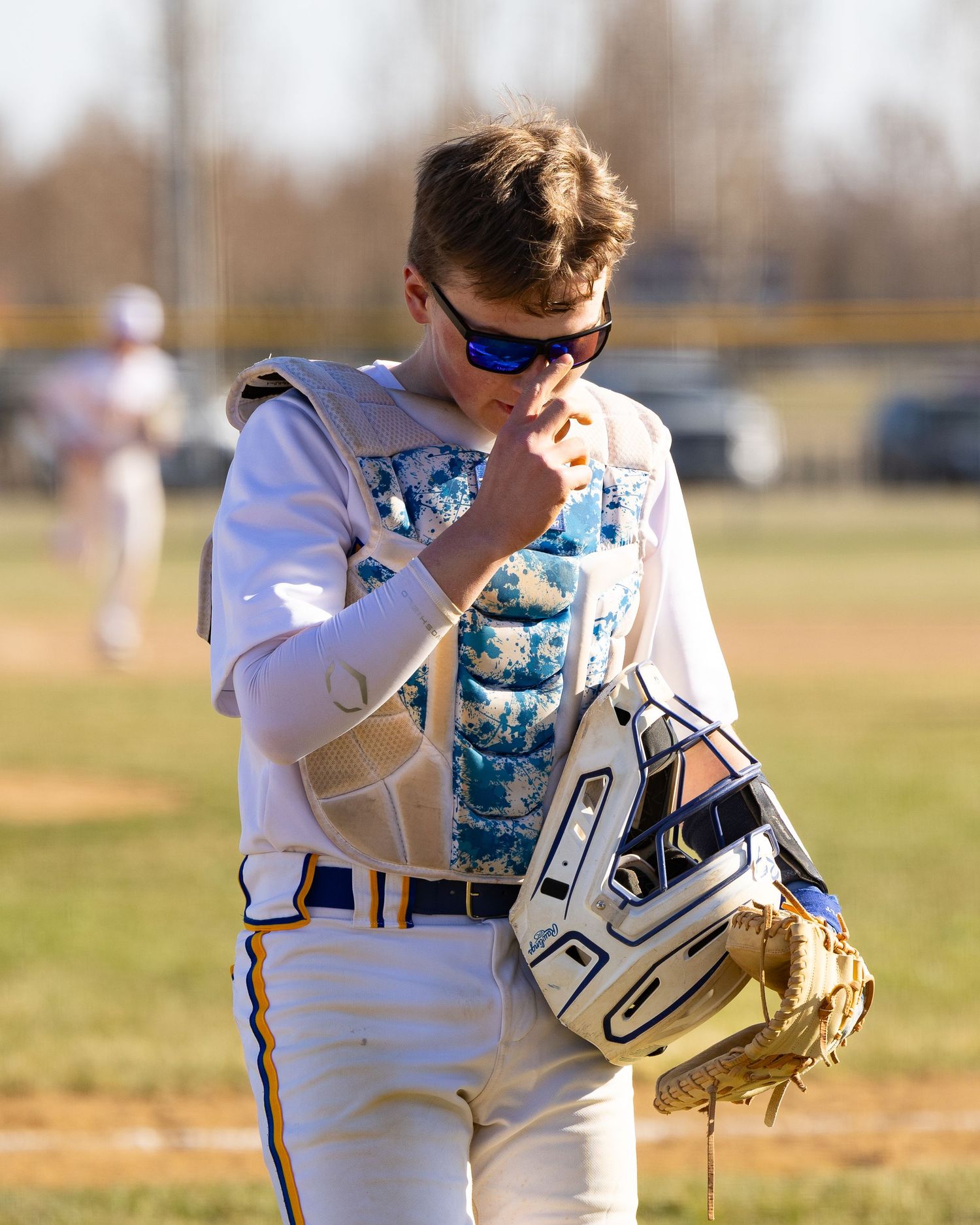 Baseball player in white uniform carries helmet and glove while walking on sunny outdoor baseball field.