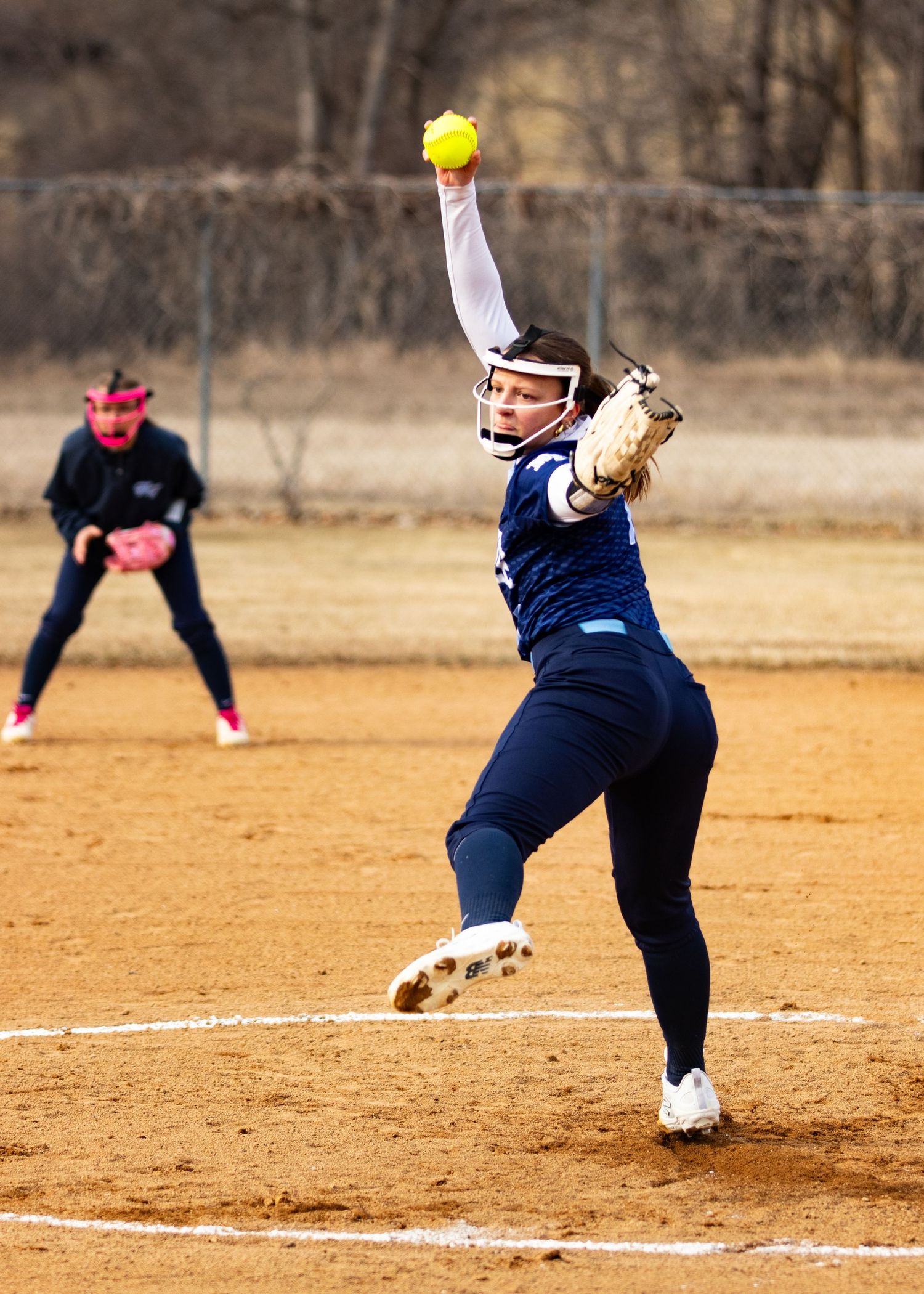 Softball pitcher in navy uniform winds up to throw yellow ball during outdoor game with fielder in background.