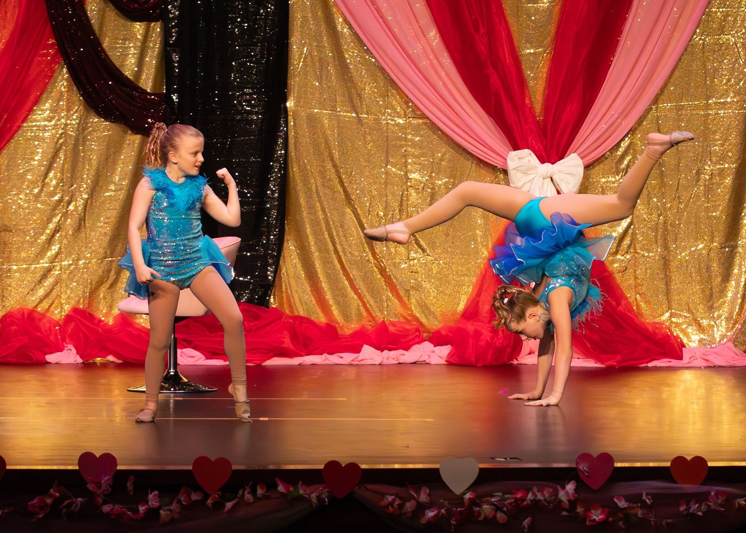 Two young dancers in turquoise costumes perform acrobatic moves on a glittering gold stage backdrop.