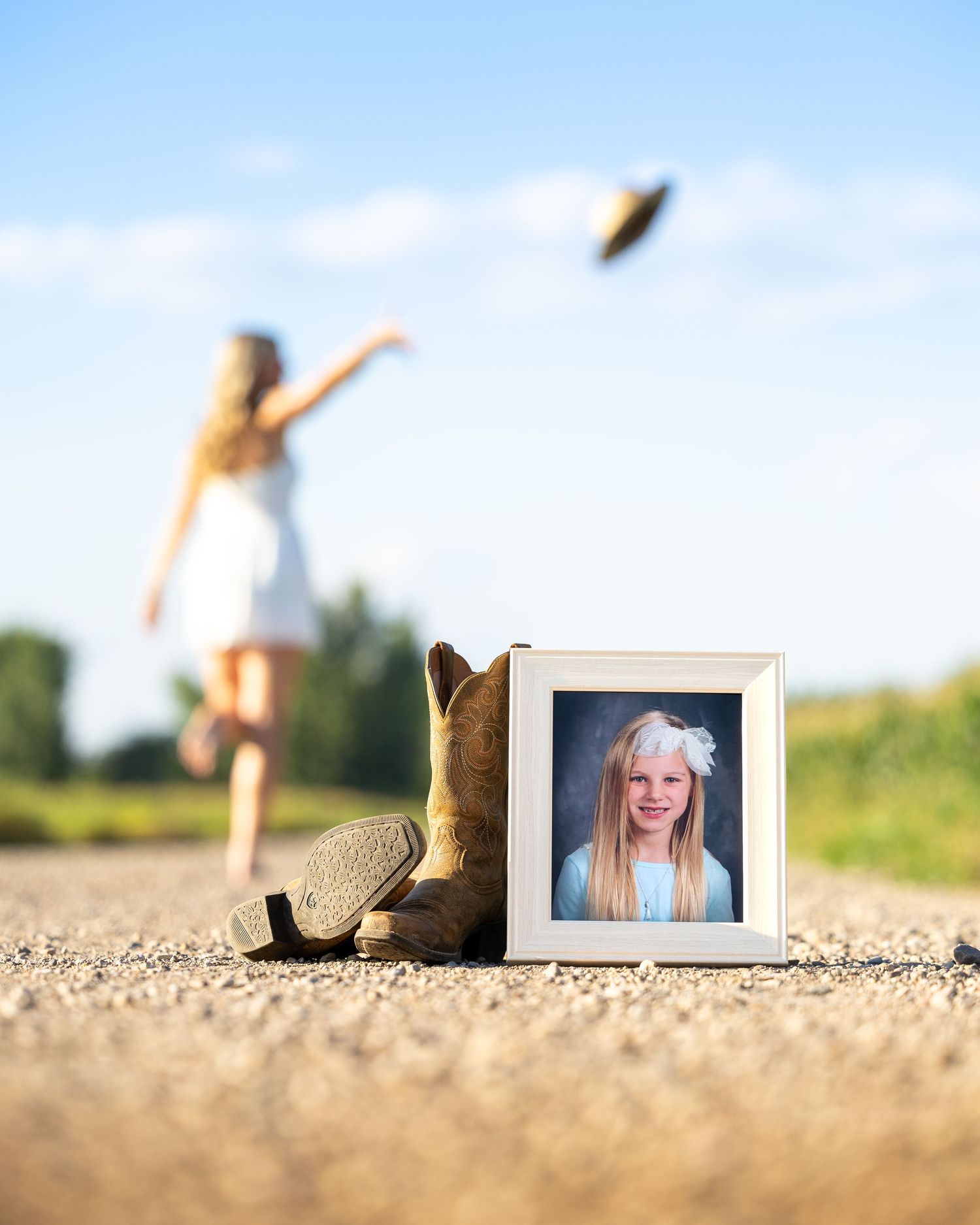 Girl throwing hat on road with framed childhood photo and cowboy boots in foreground, senior portrait concept.