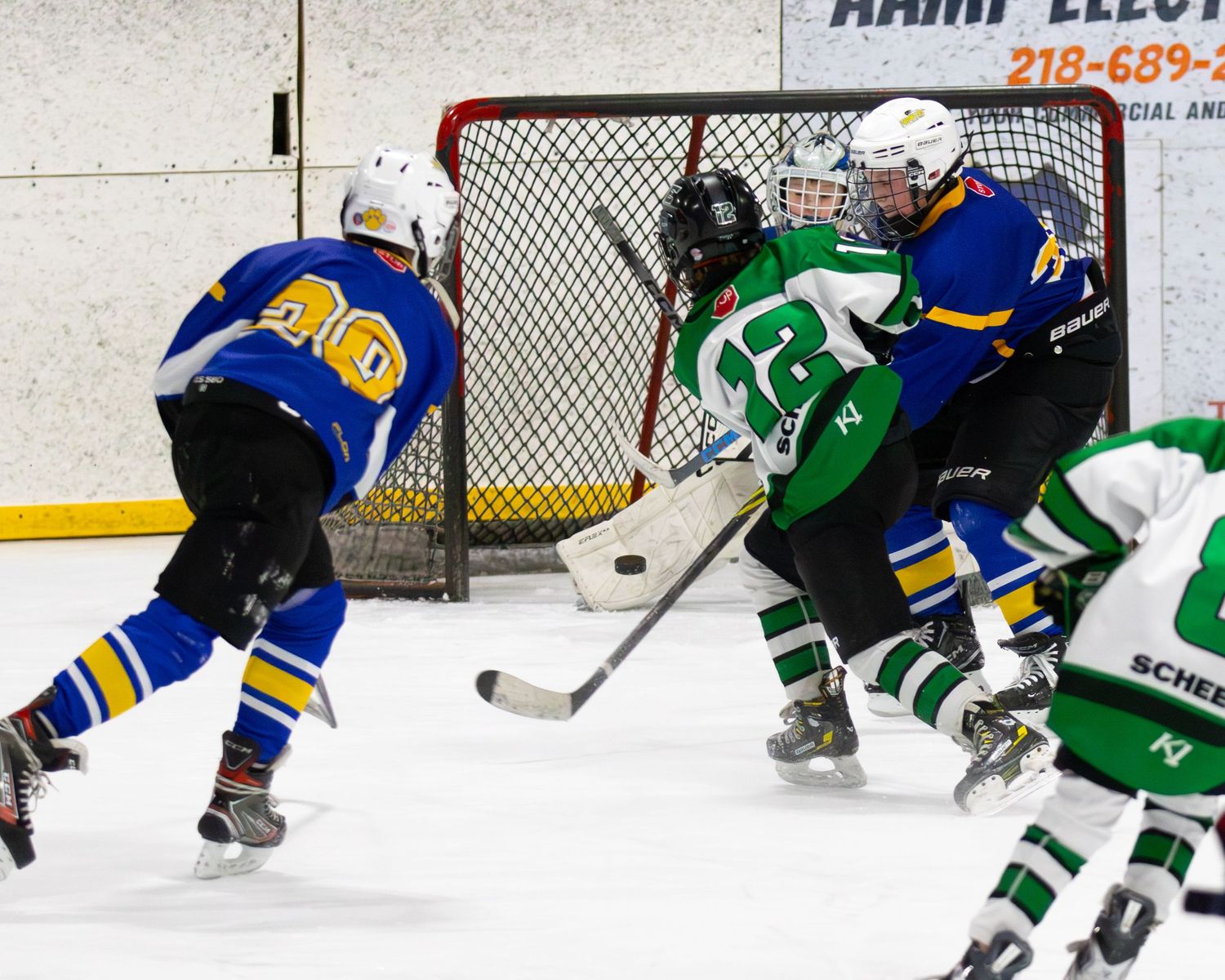 Ice hockey players in blue and green jerseys compete near the goal during an intense rink game.