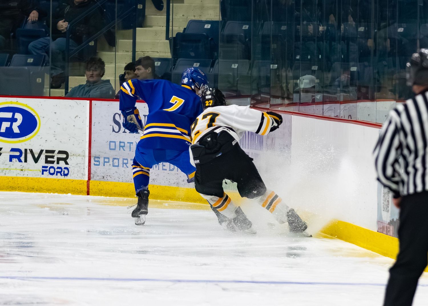 Hockey player in blue jersey number 7 battles opponent near the boards spraying ice shavings.