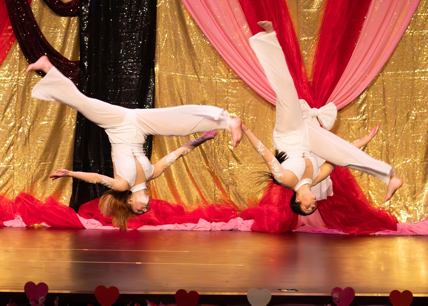 Two dancers perform synchronized horizontal flips on stage in white costumes with a gold backdrop.