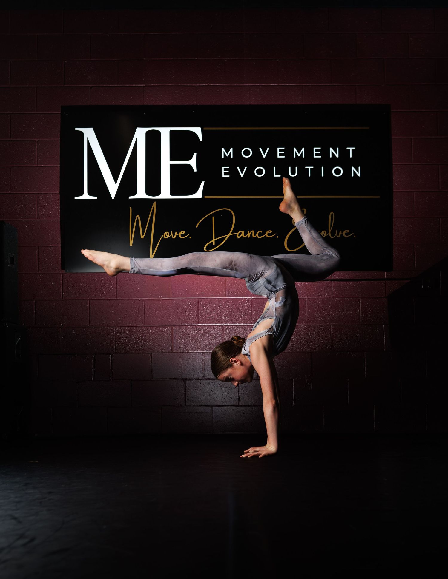 Dancer performing a one-handed handstand split in front of a Movement Evolution studio banner.