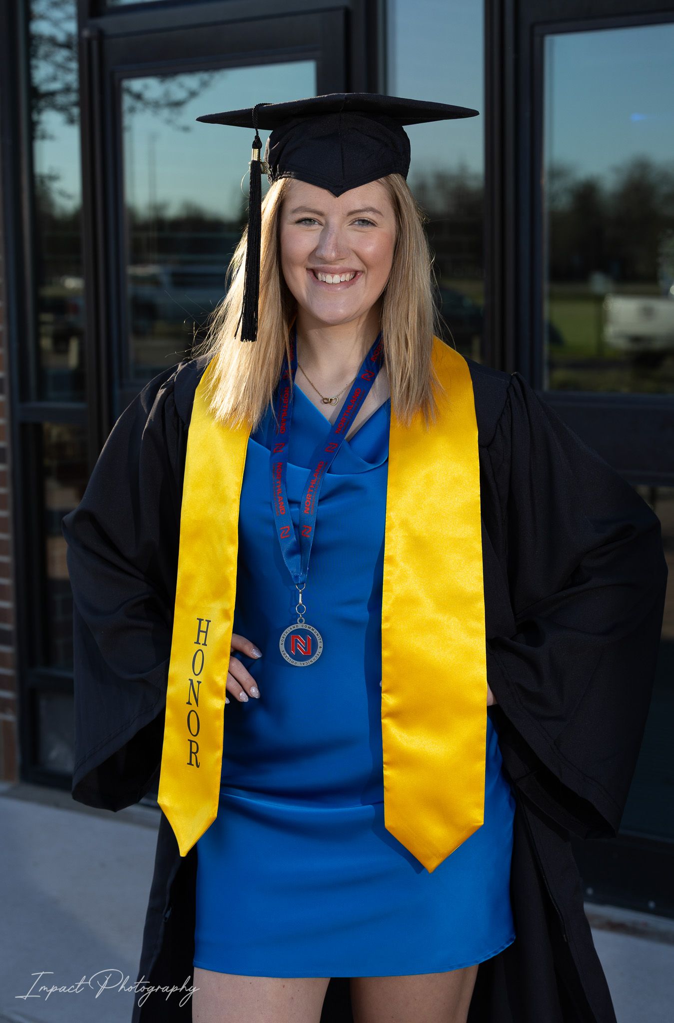 Smiling female graduate in black cap and gown with gold honor stole and blue dress posing outdoors.