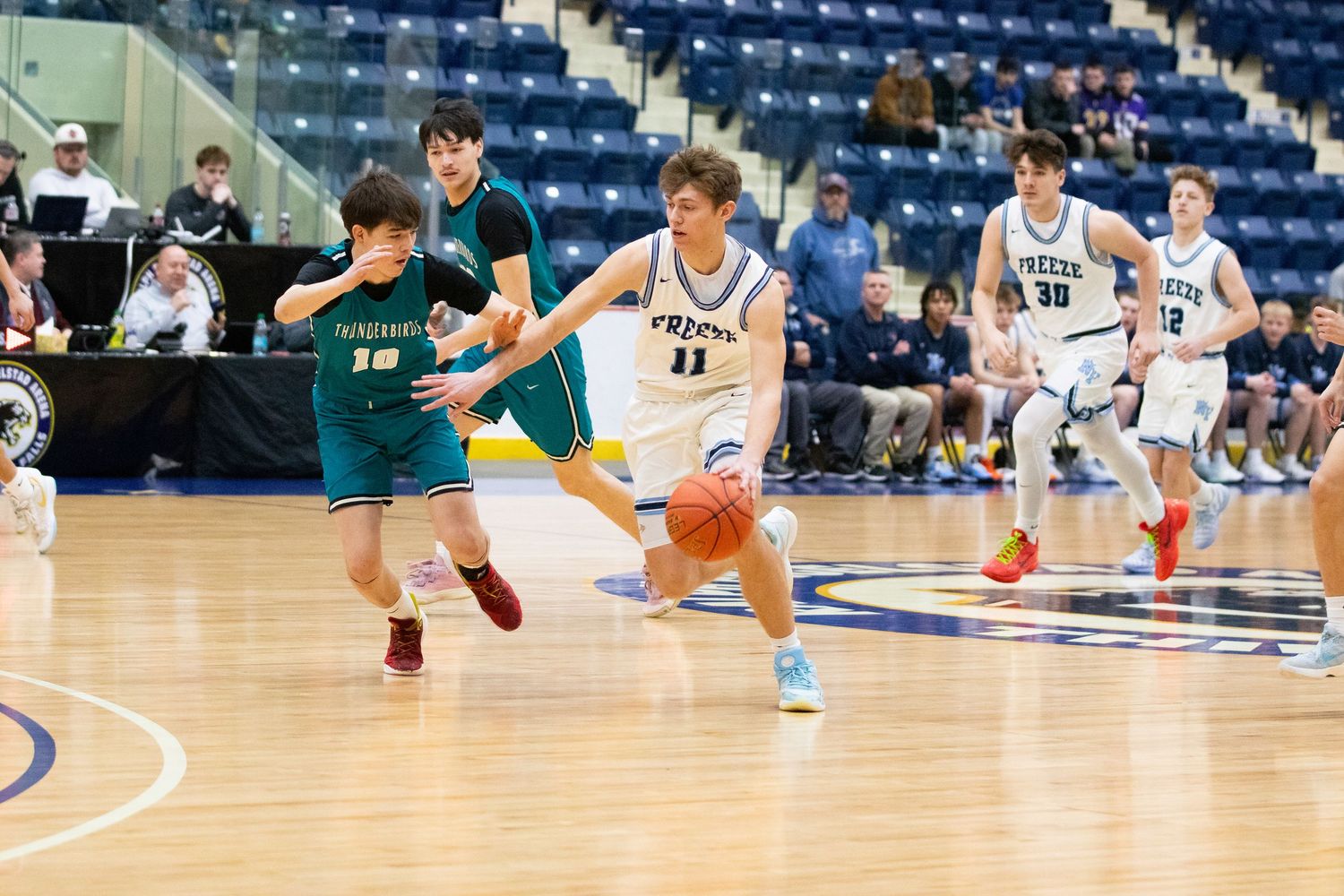 High school basketball players compete on indoor court, player #11 in white dribbles past green-uniformed defender.