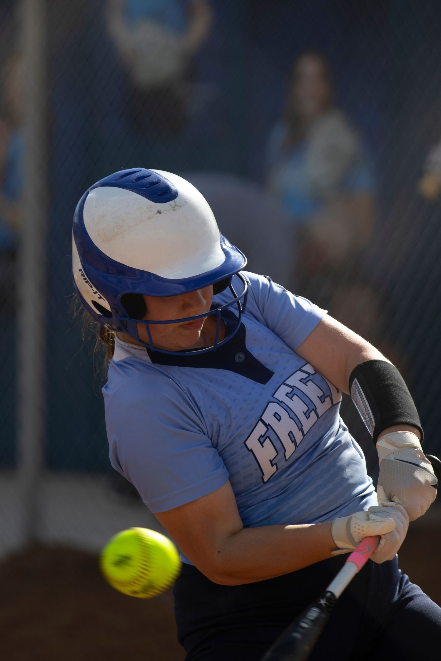 Female softball player in blue jersey and helmet makes contact with yellow ball during competitive game.