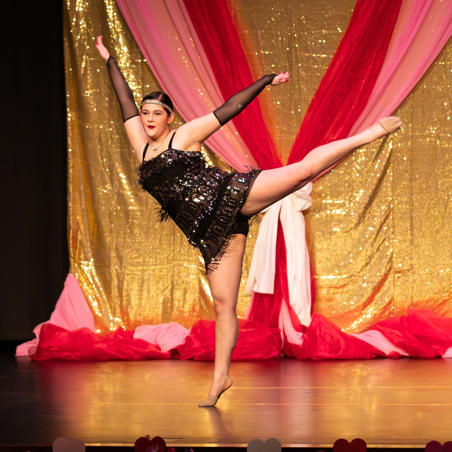 Dancer in black sequin fringe costume performs a high kick on stage with gold sequin and red drape backdrop.