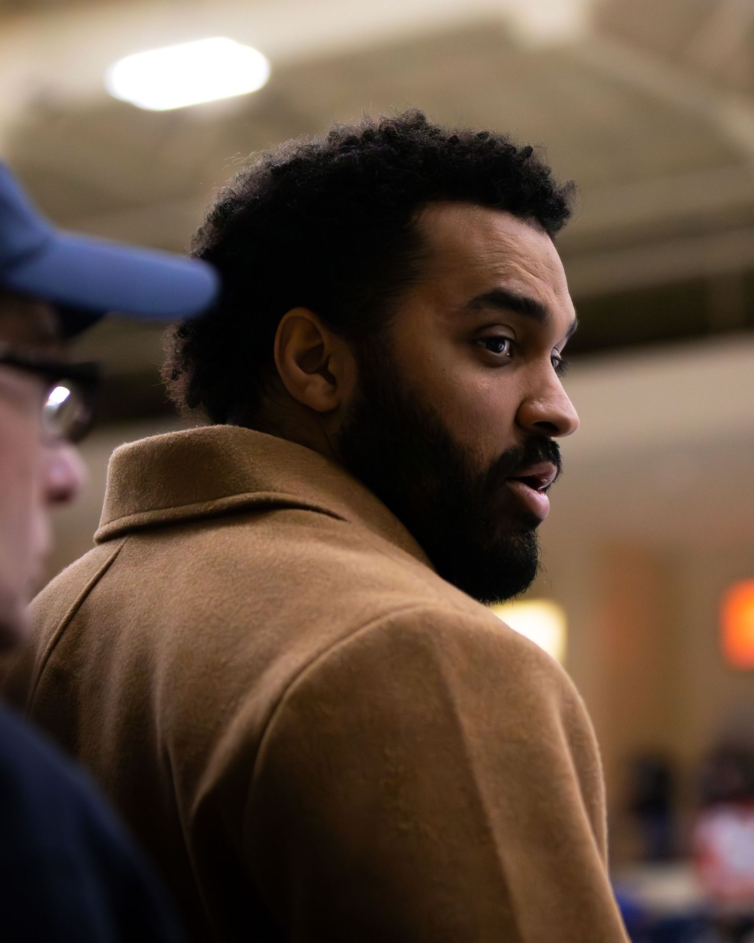 Bearded man in camel coat engaged in conversation at indoor sporting event. Hockey game.