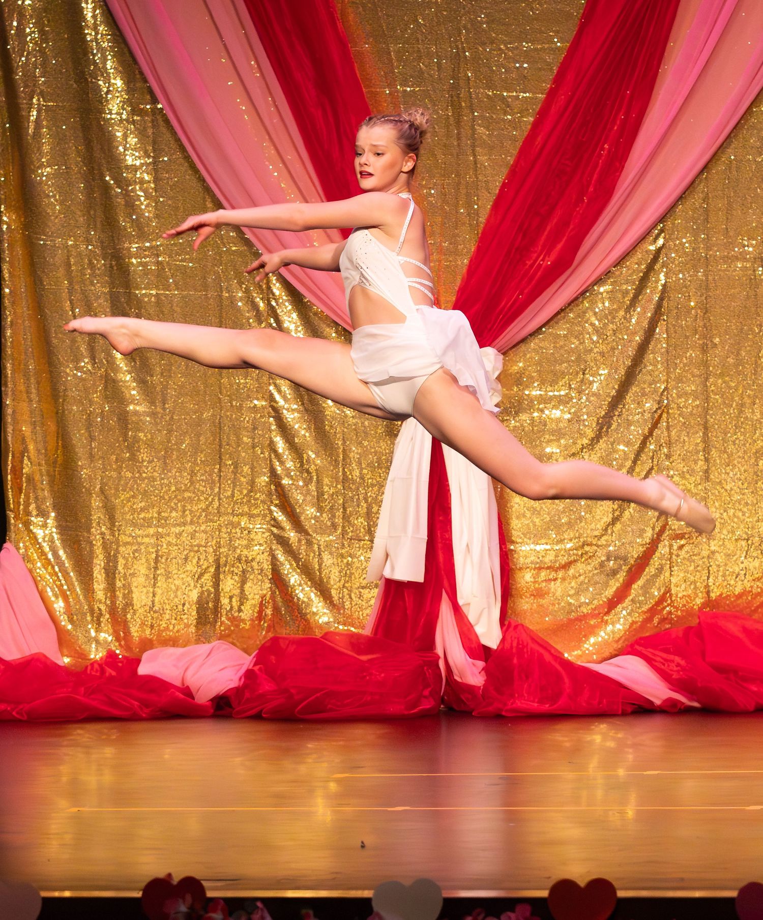 Dancer in white lyrical costume performs a graceful split leap on stage with gold sequin and red drape backdrop.