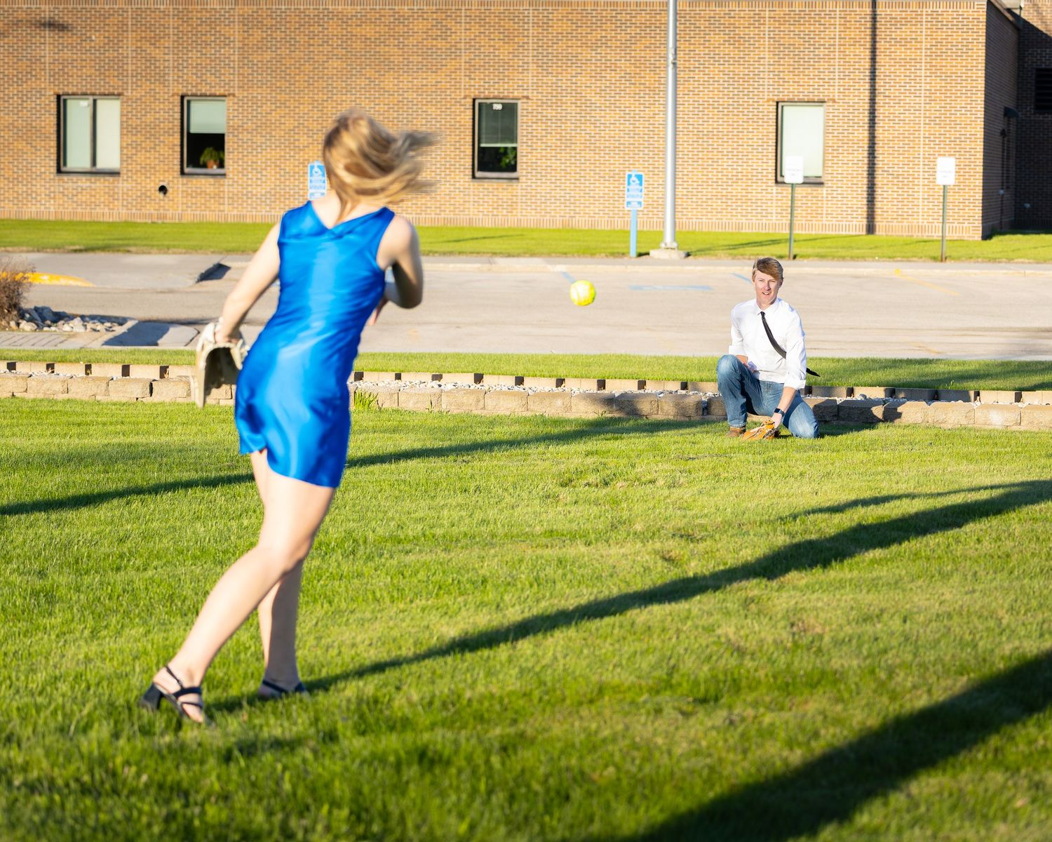 Woman in blue dress kicking a yellow balloon on green grass while a man sits watching in the background.
